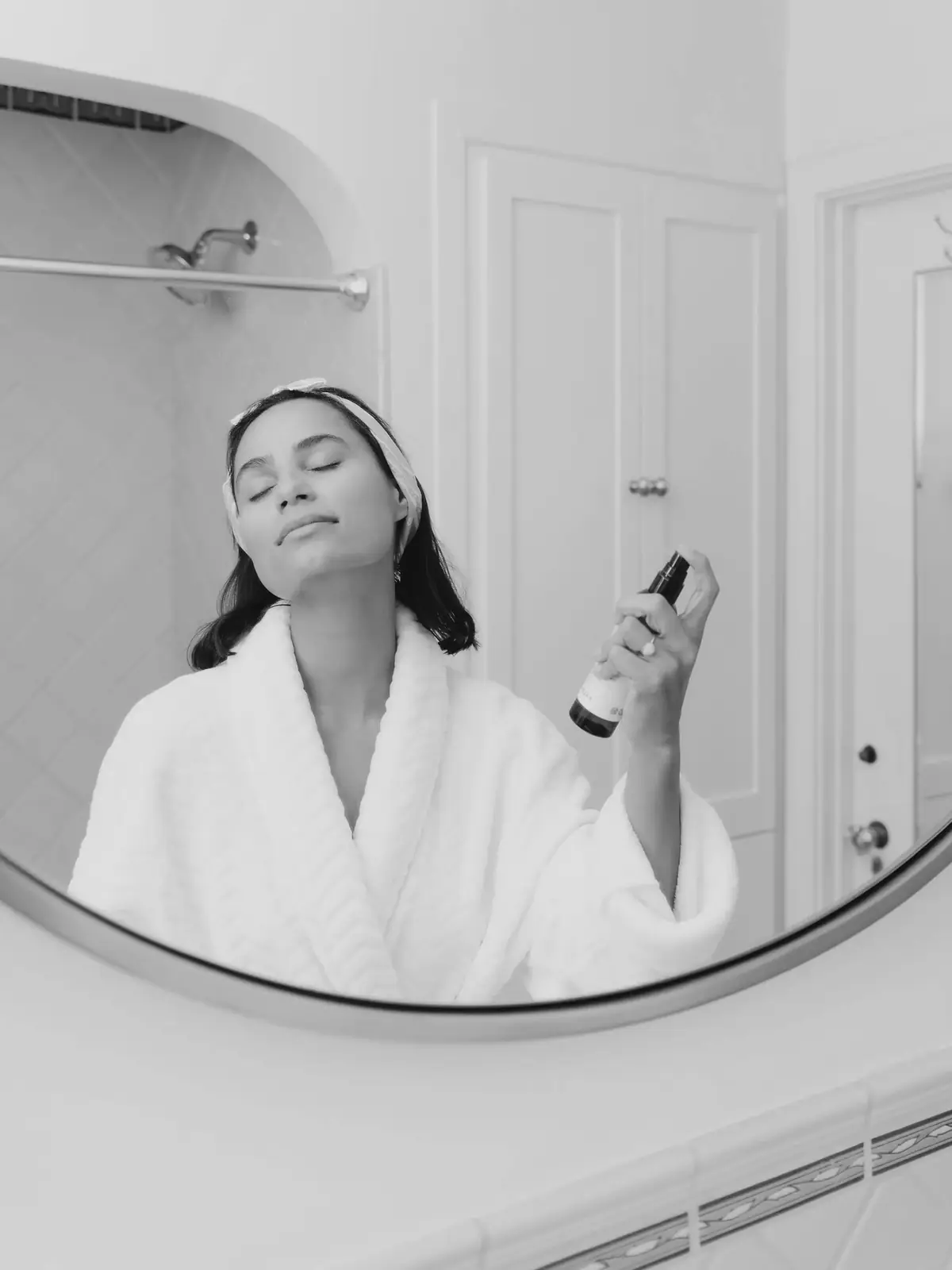 A woman in a robe misting her face in a round mirror